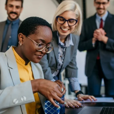 Employee is opening a gift while surrounded by her smiling team.