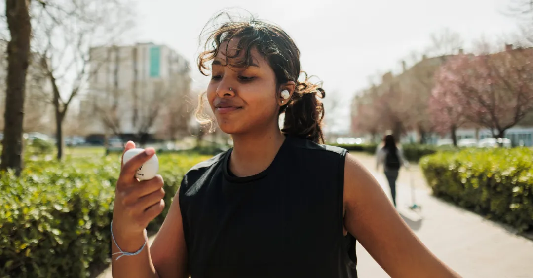 Smiling jogger wearing ear buds in a park is looking at their ear bud charger.