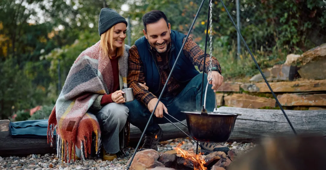 A couple cooking on a campfire. One person has a branded blanket around their shoulders.