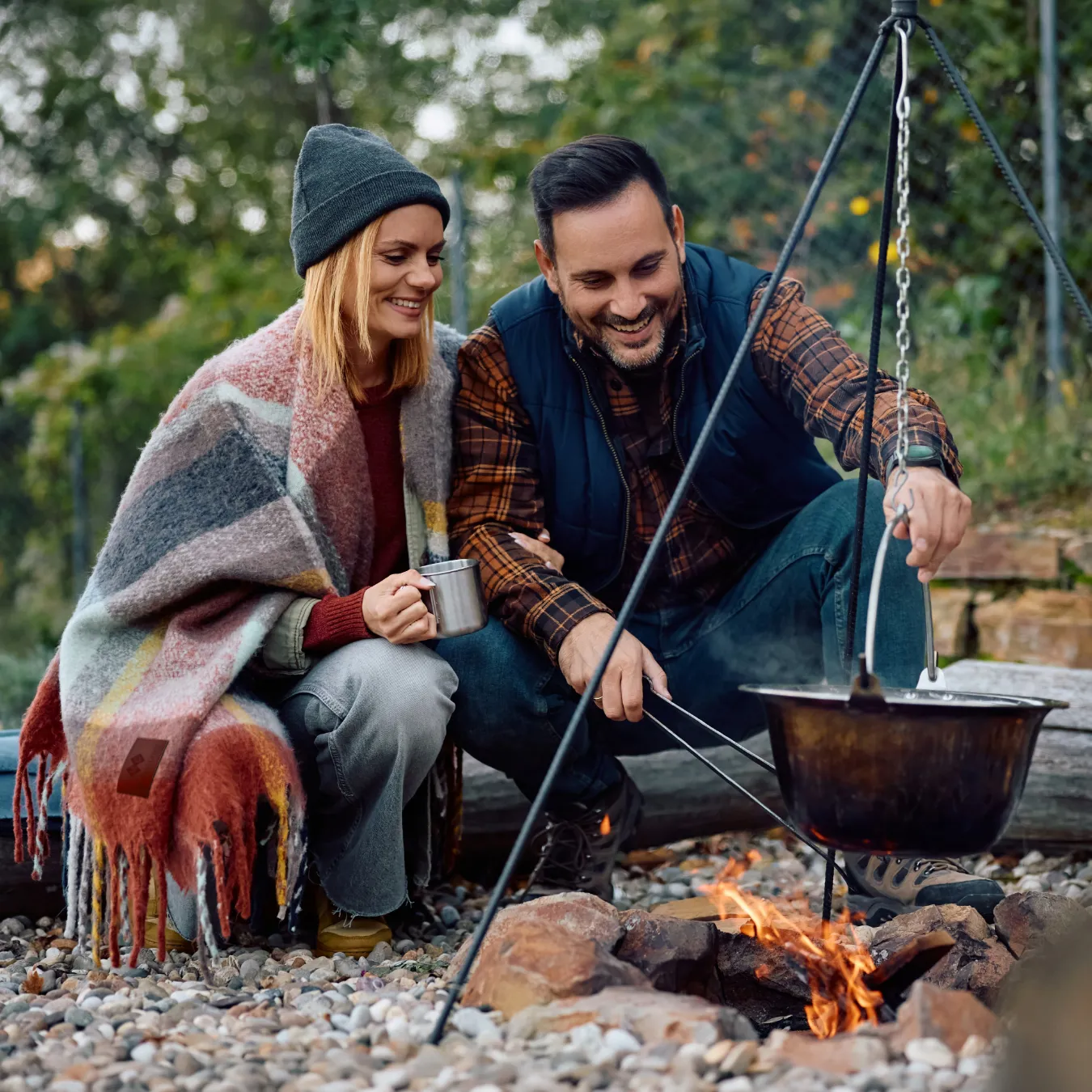 A couple cooking on a campfire. One person has a branded blanket around their shoulders.