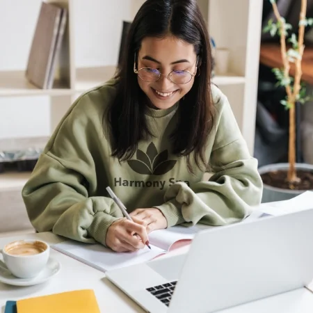 Young person in a green sweatshirt sitting at a desk in front of a laptop writing in a notebook.
