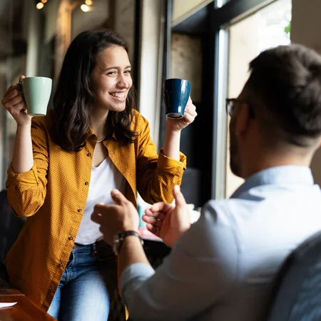 Woman sitting at a desk holding up two mugs to her coworker