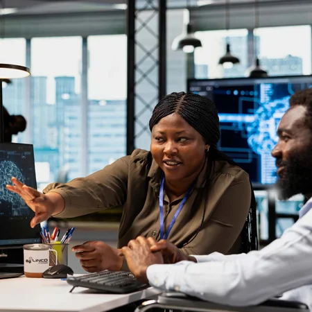 Boss explaining onboarding to new employee at desk in front of computer monitors