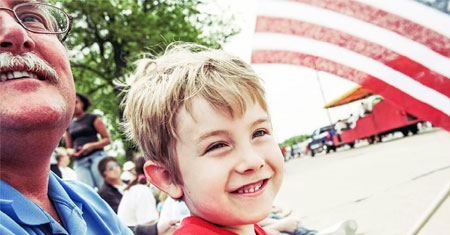 An elderly man and a young boy smile as they look ahead, with an American flag waving prominently in the background.