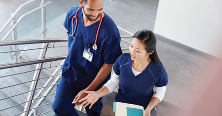 Two healthcare professionals in navy scrubs walk together, smiling and engaged in conversation.