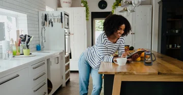 Person in standing in the kitchen looking at a tablet and drinking coffee.