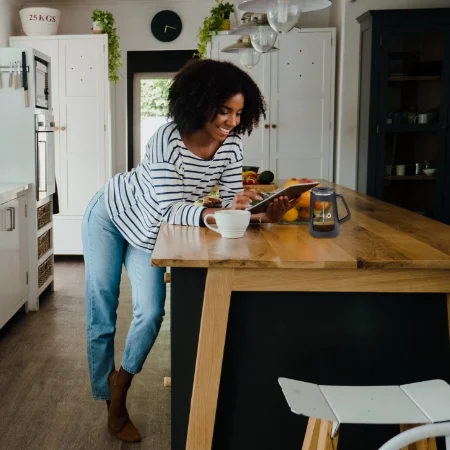 Person in standing in the kitchen looking at a tablet and drinking coffee.