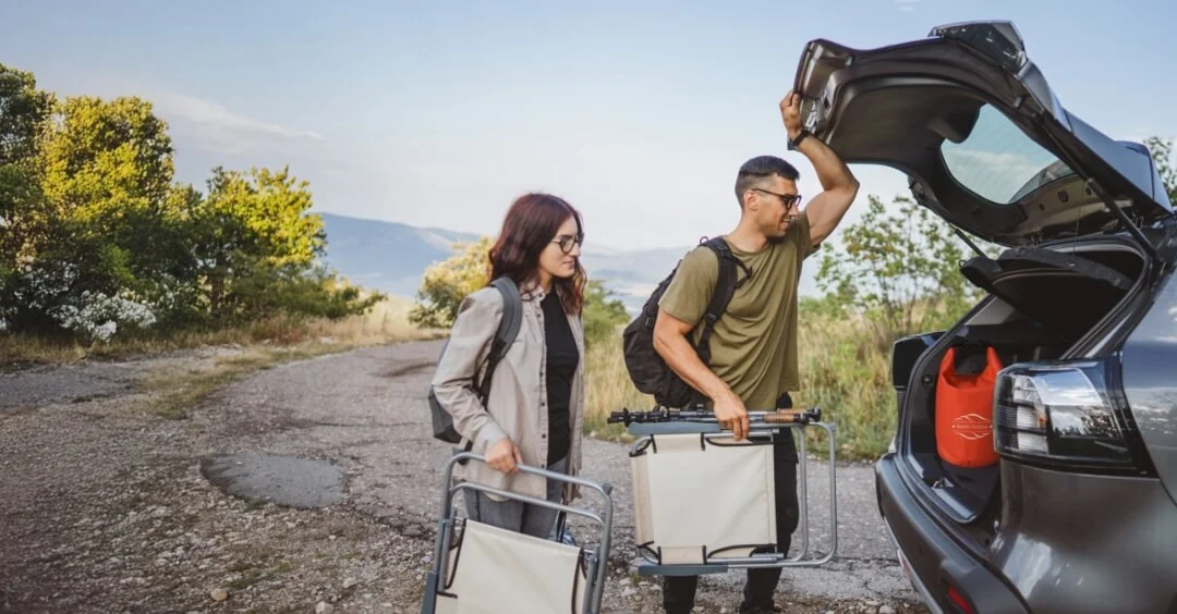 Couple loading the back of their car with chairs and camping gear. A red safety bag is sitting in the trunk.